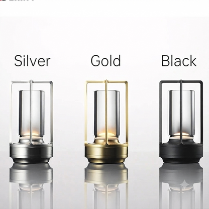 Three lantern-style table lamps shown side by side on a light gray reflective surface, labeled Silver, Gold, and Black above each variant. The lamps have clear glass bodies with metal frames and bases in silver, gold, and black.
