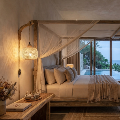 A rustic wooden four-poster bed with white and beige bedding, draped with sheer white mosquito netting, set in a cozy bedroom with a warm glowing bedside lamp, a small wooden bench, and large windows overlooking a pool and ocean.