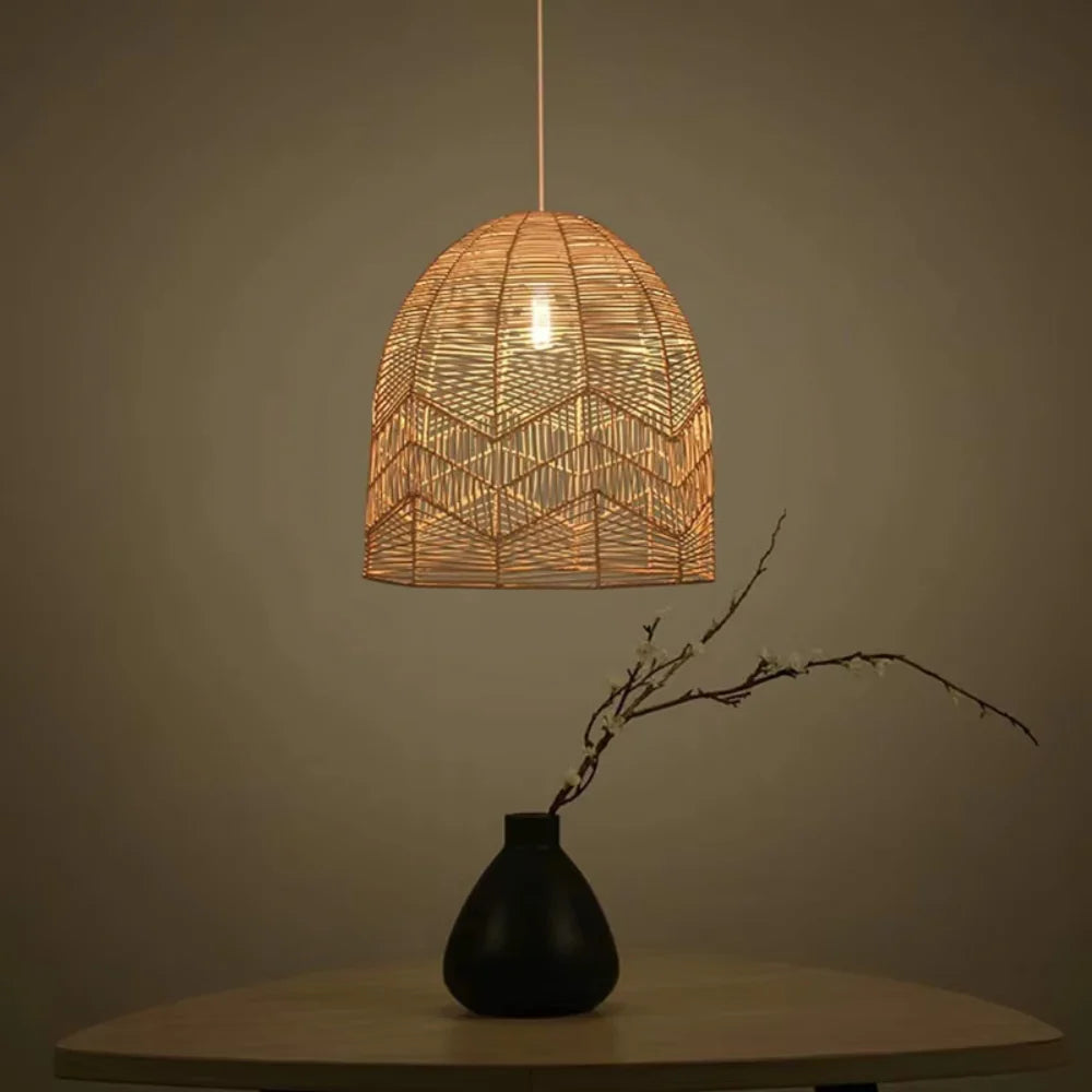 Chevron weave rattan pendant light in a bell shape hanging above a minimalist black vase on a wooden table.