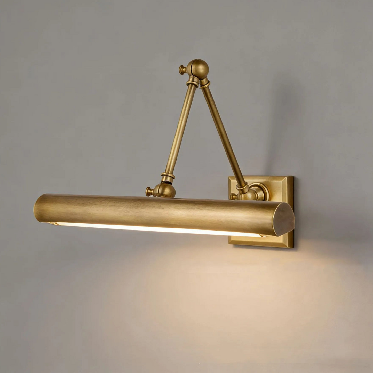 A wall-mounted cylindrical light fixture with adjustable articulated arms, finished in a warm brushed brass or gold tone, shown lit against a plain gray wall.