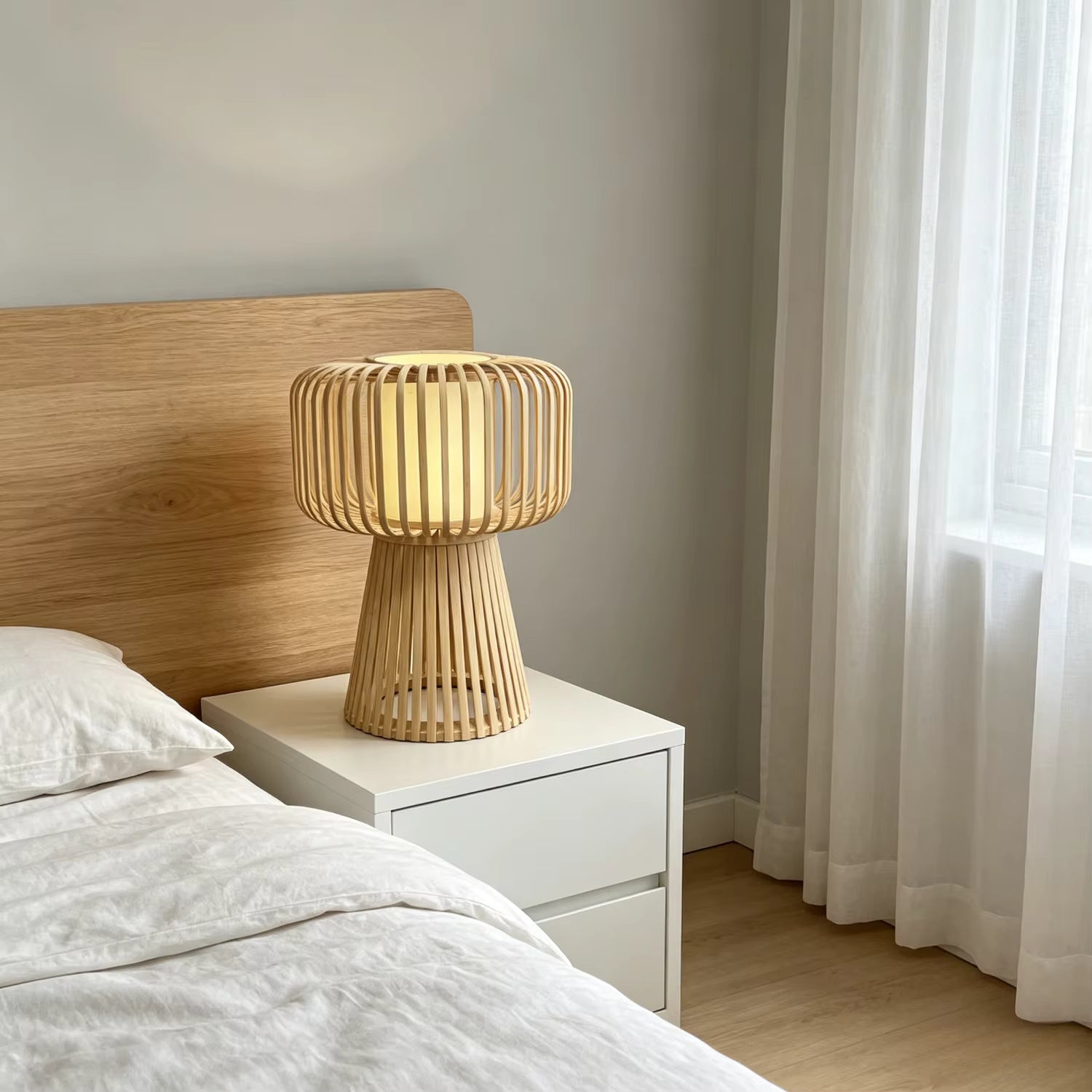 A modern bamboo slat table lamp glowing with warm light, styled on a white nightstand next to a bed with a light wood headboard in a softly lit bedroom.