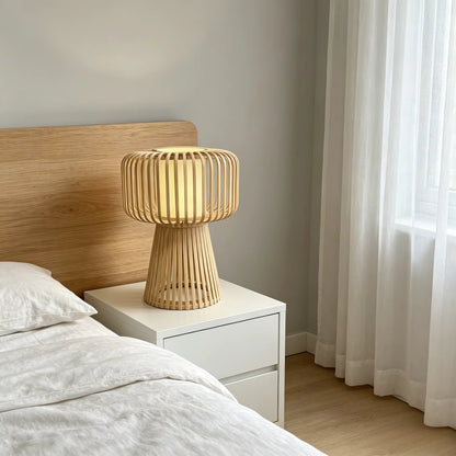 A modern bamboo slat table lamp glowing with warm light, styled on a white nightstand next to a bed with a light wood headboard in a softly lit bedroom.