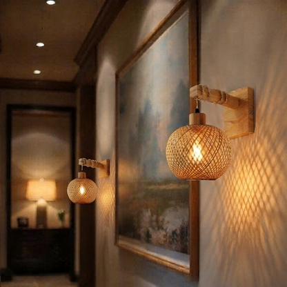 A warm, bohemian-style wall sconce with a woven rattan globe and a light wood wall mount, shown illuminated in a hallway next to a framed painting.