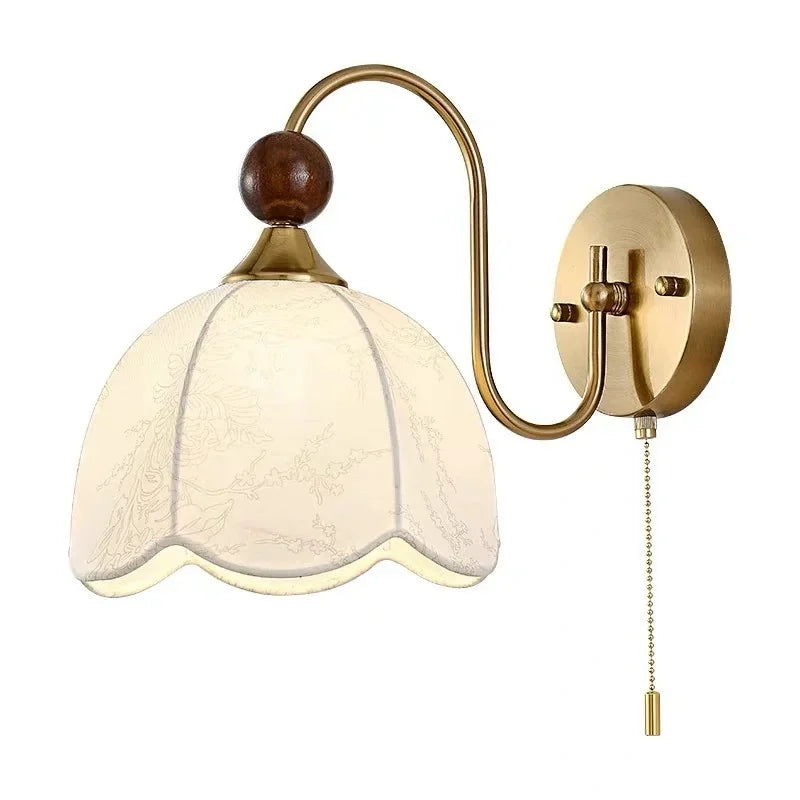 A vintage-style wall sconce light fixture with a curved brass arm, a round wall mount, a bell-shaped cream-colored glass shade with a subtle floral pattern, a dark brown wooden finial, and a pull chain.