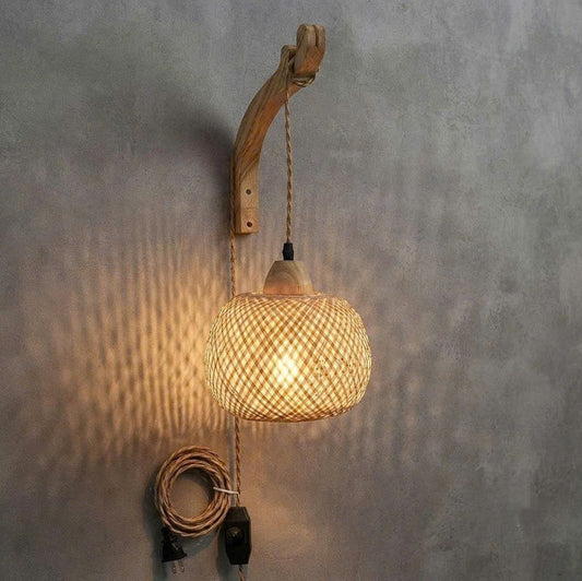 A rustic wall-mounted pendant light with a curved natural wood backplate, a woven rattan shade, and a hanging cord with a plug, shown against a textured gray wall.