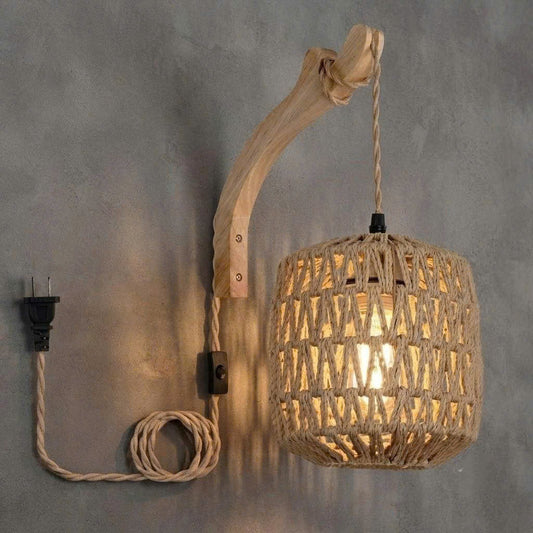 A rustic wall-mounted pendant light with a natural wood curved arm, a woven rope shade, and a coiled cord with a black plug, shown against a textured gray wall.