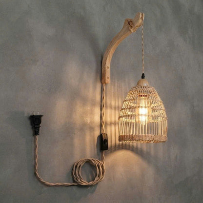 A rustic wall-mounted pendant light with a curved natural wood branch arm, a woven rattan birdcage shade, and a coiled beige cord with a black plug, shown against a textured gray wall.