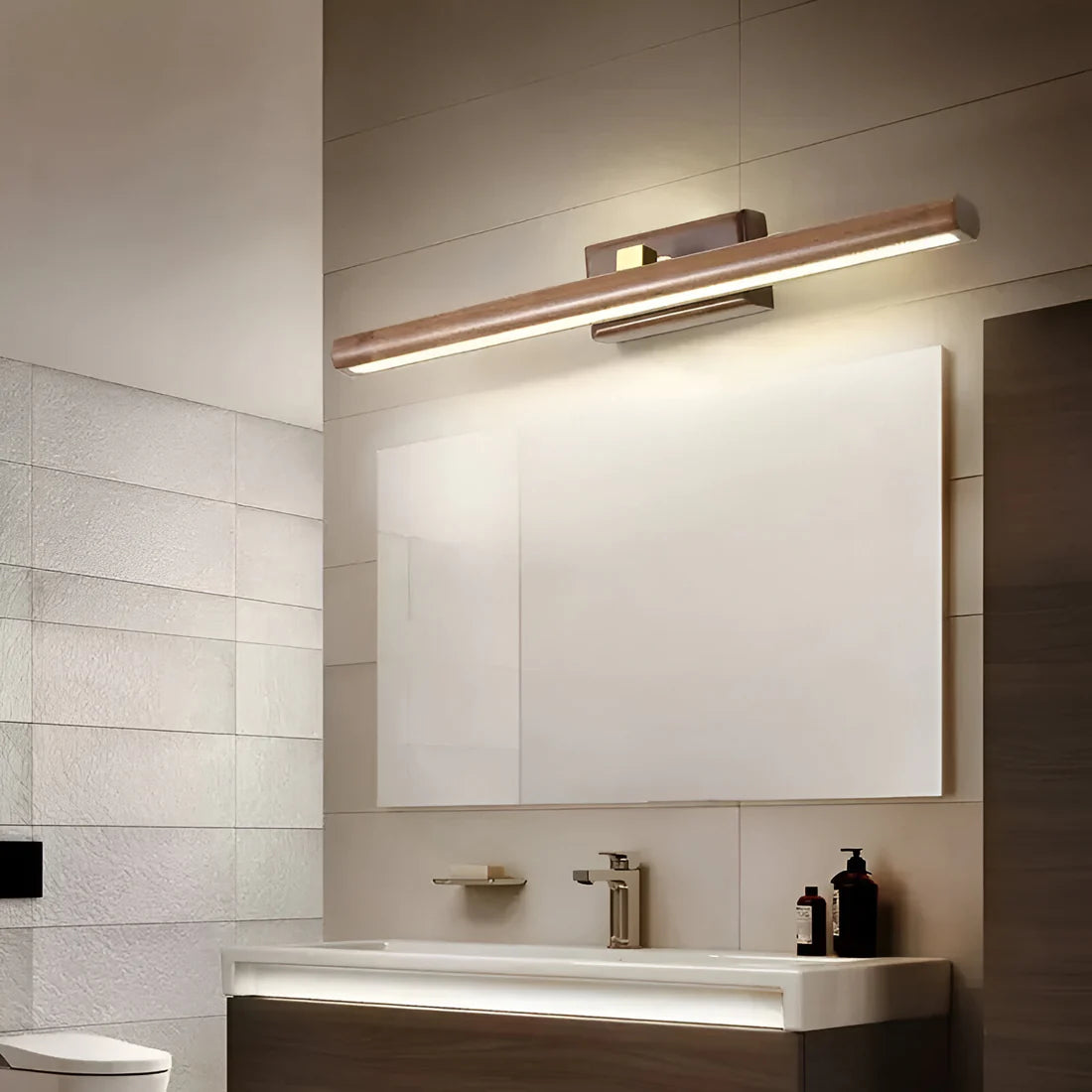 A modern wall-mounted bathroom light fixture with a brushed brass-toned rectangular bar and integrated LED lighting, installed above a white sink and mirror in a contemporary bathroom setting.