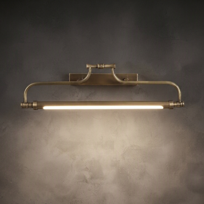 A vintage-style industrial wall sconce with a brushed brass finish, featuring a horizontal bar with a glowing light tube, mounted on a textured dark gray wall.