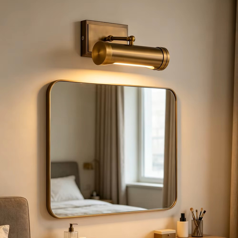 A cylindrical wall-mounted light fixture in a warm brass or bronze finish, mounted above a rectangular mirror on a beige wall. The light is turned on, casting a warm glow downward, with a softly blurred bedroom scene reflected in the mirror.