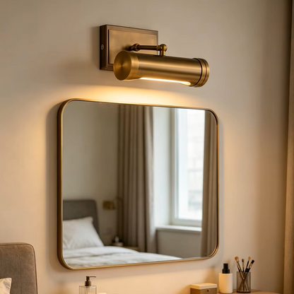 A cylindrical wall-mounted light fixture in a warm brass or bronze finish, mounted above a rectangular mirror on a beige wall. The light is turned on, casting a warm glow downward, with a softly blurred bedroom scene reflected in the mirror.