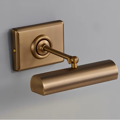 A modern wall-mounted light fixture with a cylindrical shade and rectangular backplate in a brushed brass or bronze finish, shown against a plain gray wall.