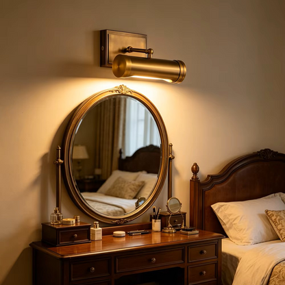 A brass wall-mounted cylindrical sconce with a warm glowing light, mounted above an ornate round vanity mirror on a bedroom wall.
