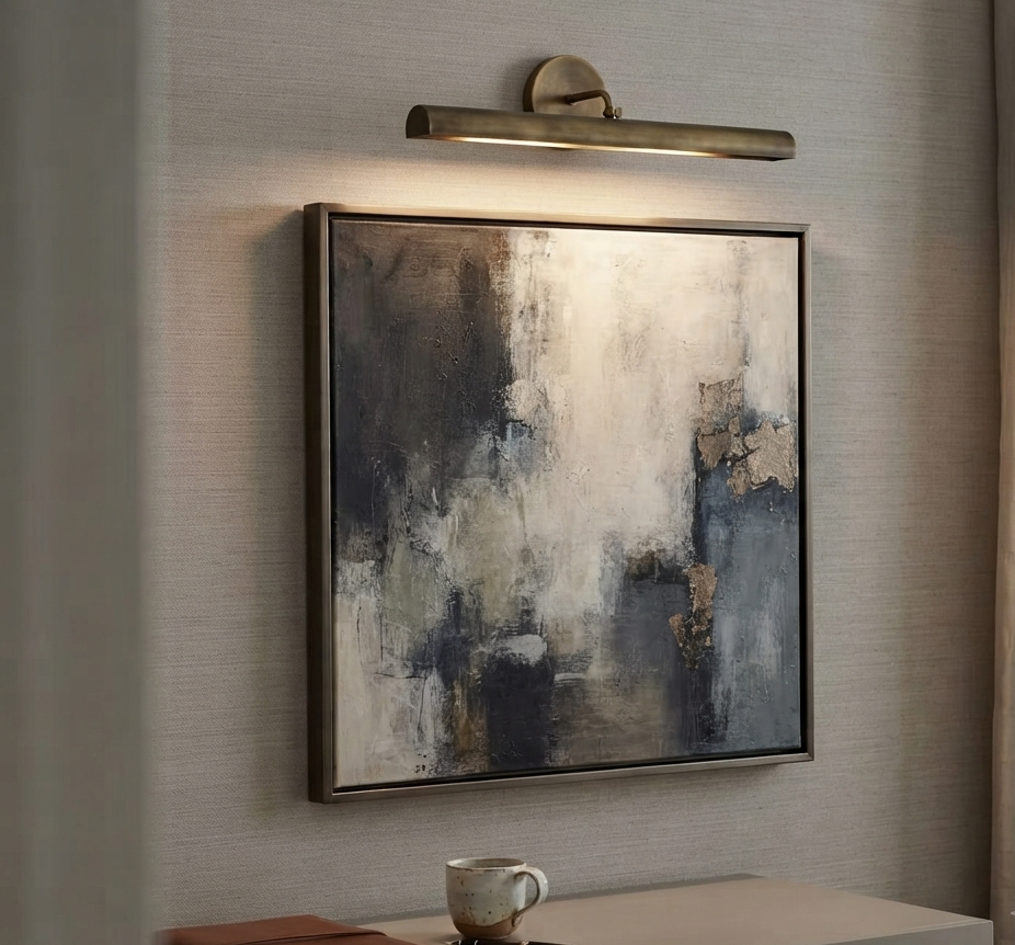 A modern wall-mounted light bar with a warm brass finish illuminates a square abstract canvas painting hanging on a textured beige wall above a console table with a mug and notebook.