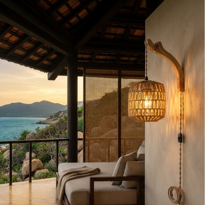 A warm, rustic outdoor lounge chair with neutral cushions sits on a covered patio overlooking a scenic ocean and mountain view at sunset. A woven pendant light hangs on the right against a textured wall.
