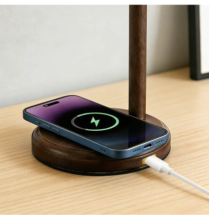 A dark brown wooden dual-device wireless charging stand on a light wood desk, with a smartphone resting on the top level and a white charging cable plugged into the bottom level.