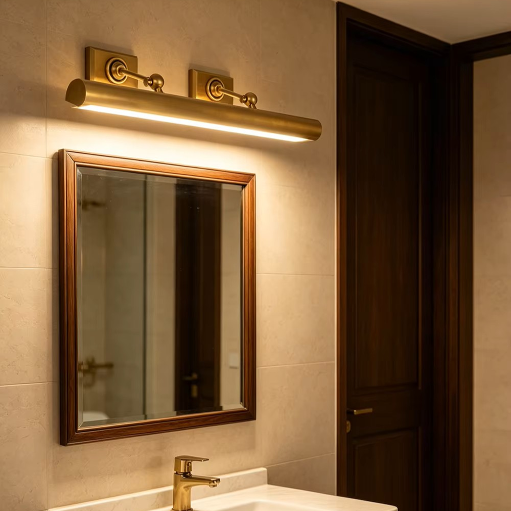A modern cylindrical wall light fixture in a warm brass/gold finish mounted above a bathroom mirror with a dark wood frame, installed in a beige tiled wall next to a dark wood door.