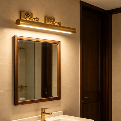 A modern cylindrical wall light fixture in a warm brass/gold finish mounted above a bathroom mirror with a dark wood frame, installed in a beige tiled wall next to a dark wood door.