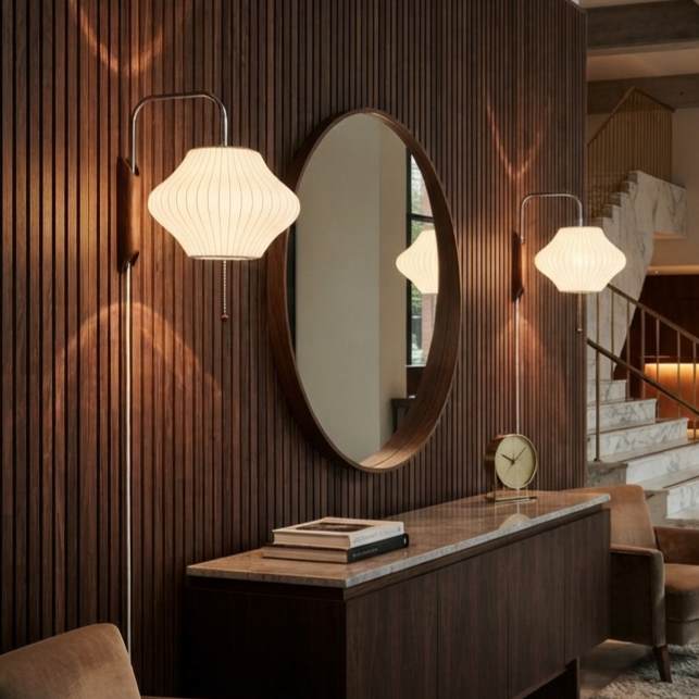 A modern interior scene with a dark wood slatted wall, an oval wall mirror, two lit floor sconces with pleated white shades, a console table, books, and a small clock. No person or animal is present, but this is still not a clean isolated product shot because it is shown installed in a real room environment, so subject_type is Lifestyle.
