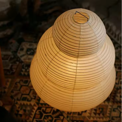 A warm white paper lantern-style lamp with a tiered, ribbed design, glowing with a soft yellow light in an indoor setting.