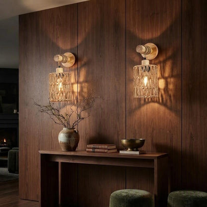 Two matching wall sconces with warm glowing bulbs and textured cylindrical shades, mounted on a dark wood-paneled wall above a console table in a styled interior setting.