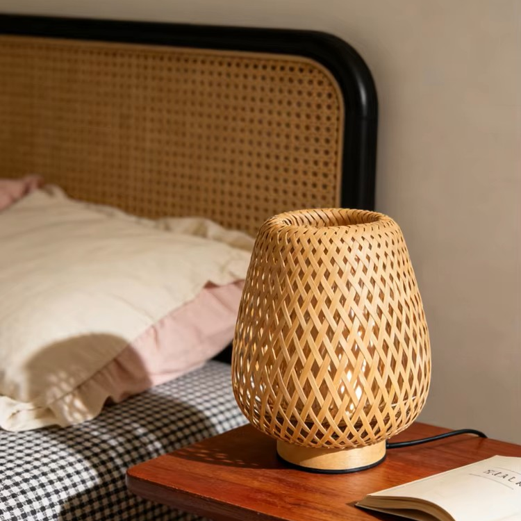 A woven bamboo table lamp with a diamond-patterned lattice design sits on a wooden bedside table next to a bed with a cane headboard.