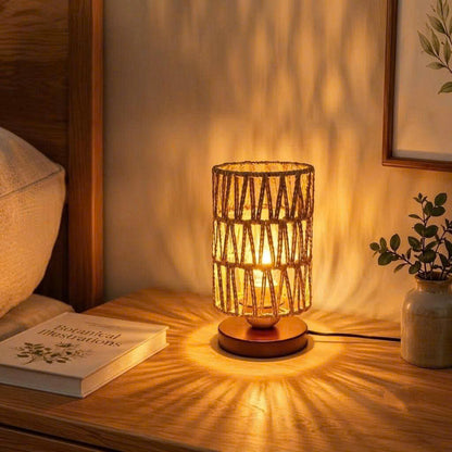 A warm-toned table lamp with a woven cylindrical shade and wooden base sits on a wooden bedside table, casting a starburst pattern of light onto the wall. The scene includes a pillow, a book, and a small potted plant in a cozy bedroom setting.
