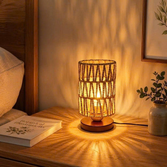 A warm-toned table lamp with a woven cylindrical shade and wooden base sits on a wooden bedside table, casting a starburst pattern of light onto the wall. The scene includes a pillow, a book, and a small potted plant in a cozy bedroom setting.