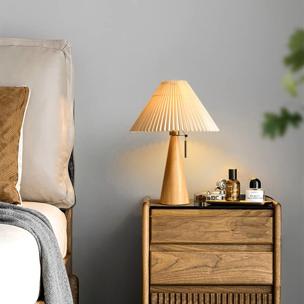 A warm-toned table lamp with a pleated beige shade and a light wood base sits on a wooden nightstand in a bedroom setting next to a bed.