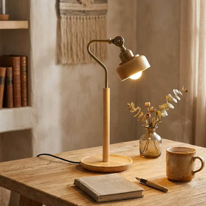 A warm, rustic table lamp with a light wood base and stem, a curved metal neck, and a beige shade glowing with a warm light, styled on a wooden desk next to a mug, notebook, and small vase.