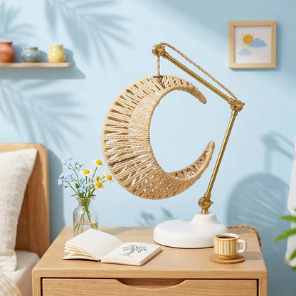 A crescent moon-shaped table lamp with a woven natural rattan shade and a gold-finished adjustable arm on a white base, styled on a wooden nightstand in a bedroom setting.