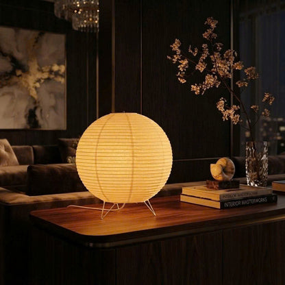 A glowing spherical paper lantern table lamp sits on a wooden console table in a dimly lit living room setting, with books, a vase of branches, and dark wood furniture in the background.