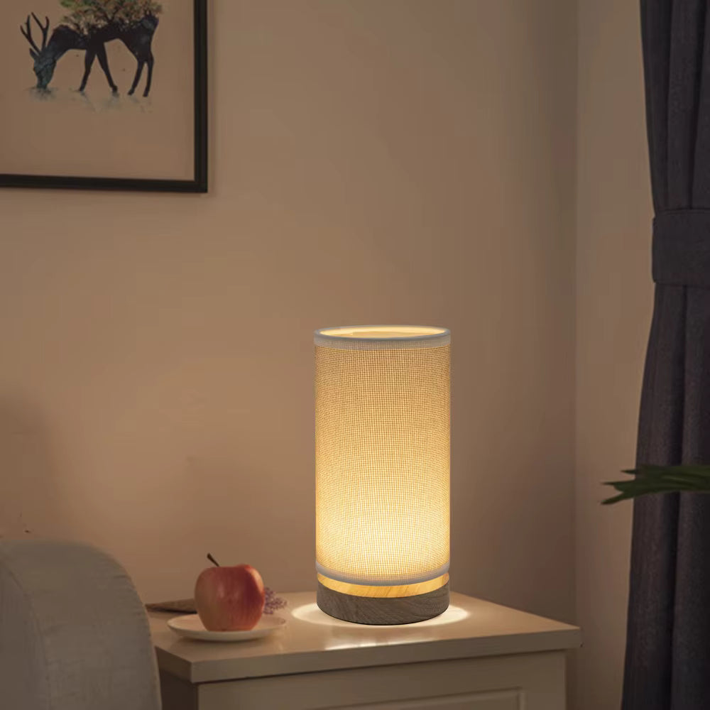 A cylindrical table lamp with a warm glowing fabric shade and a textured gray base, placed on a bedside table in a softly lit room.