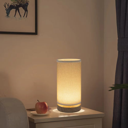 A cylindrical table lamp with a warm glowing fabric shade and a textured gray base, placed on a bedside table in a softly lit room.