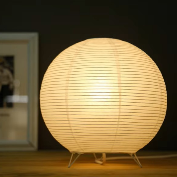 A glowing spherical table lamp made of horizontal ribbed paper or fabric, standing on thin metal legs on a wooden surface against a dark indoor background.