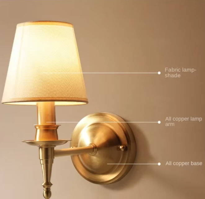 A wall-mounted brass sconce with a warm glowing fabric lampshade, shown against a beige wall with callout lines labeling the fabric lampshade, copper lamp arm, and copper base.