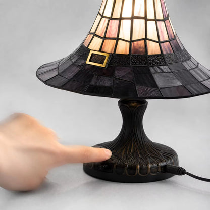 A stained-glass table lamp with a black sculpted base and a conical shade made of pink, purple, and black glass panels, shown with a hand pressing the base to turn it on.