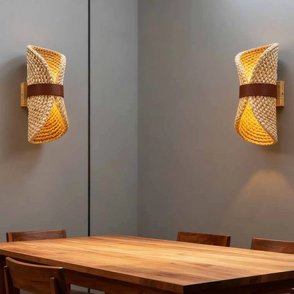 Two modern wall sconces with woven natural-fiber shades and brown leather-like bands, mounted on gray walls above a wooden dining table with chairs.