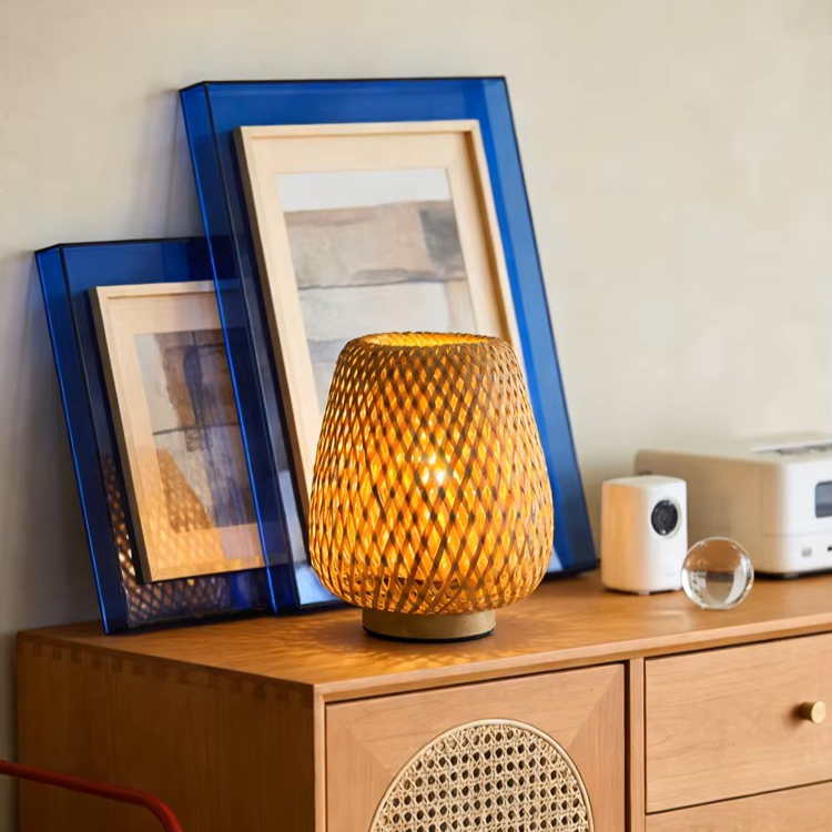 A lit woven rattan table lamp with a diamond-patterned shade sits on a wooden cabinet in a styled room setting, with blue-framed art prints and a white speaker in the background.