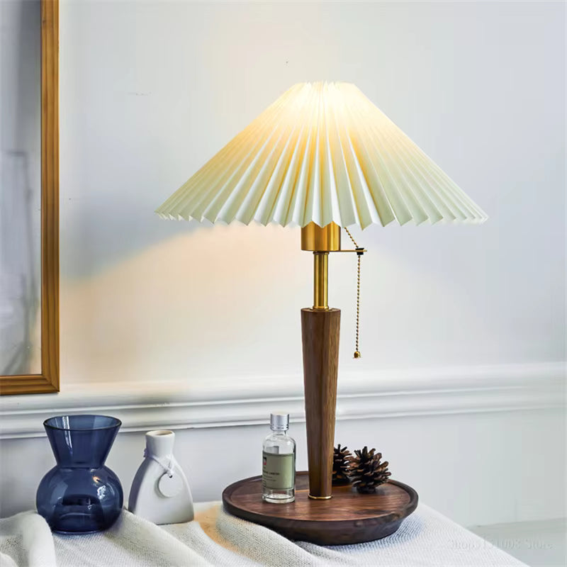 A table lamp with a pleated cream shade, a warm brass-toned neck, and a dark wood base on a round wooden tray, styled on a table with a blue glass vase, a small white ceramic object, and a bottle in a home interior setting.