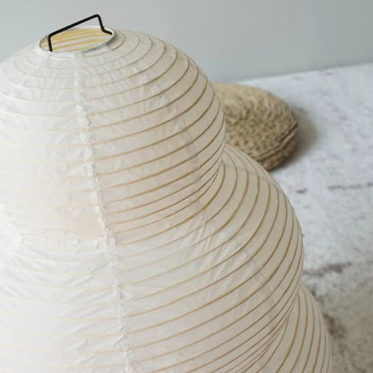 A close-up of a cream-colored paper lantern with a ribbed paper structure and a black metal handle, set against a light gray background with a woven mat prop.