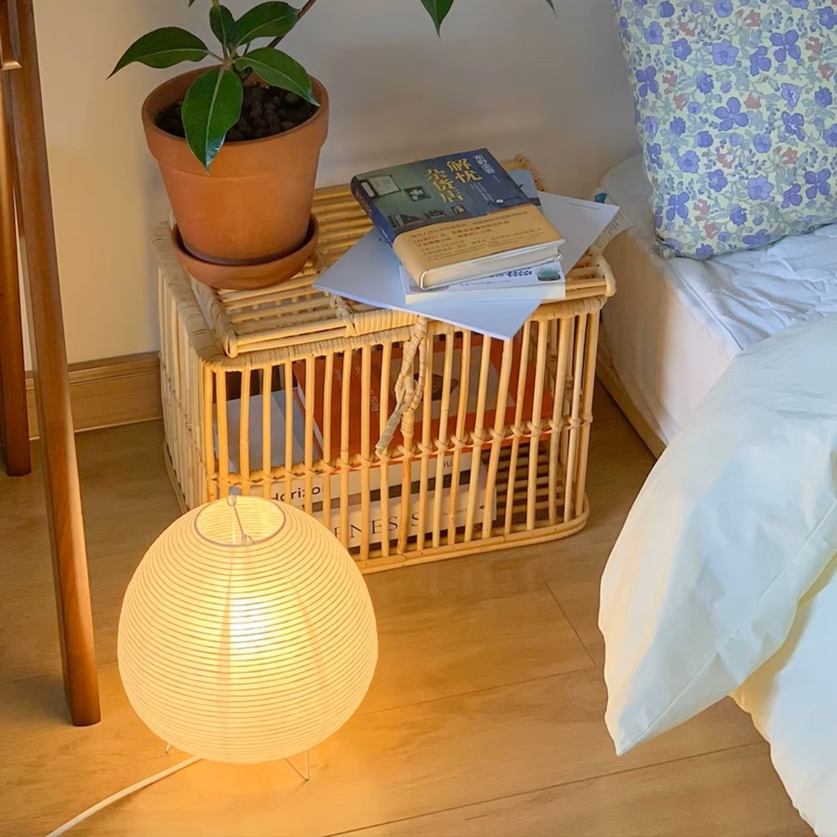 A warm, cozy bedroom corner featuring a glowing white ribbed paper lantern lamp on a light wood floor, next to a rattan storage crate holding books and a potted plant, with a bed and floral pillow in the background.