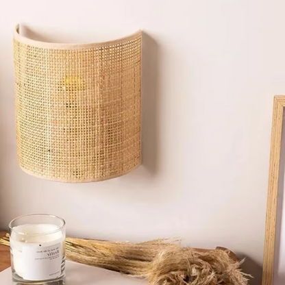 A curved wall-mounted lampshade made of natural woven rattan or cane, shown hanging on a light beige wall above a tabletop with a candle and dried grasses.