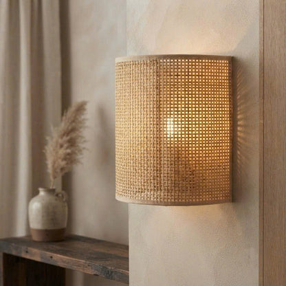A cylindrical wall sconce with a woven natural rattan shade, illuminated from within to cast a warm golden glow against a textured beige wall.