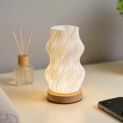 A glowing ceramic table lamp with a wavy, ribbed white shade and a round natural wood base, set on a light-colored tabletop next to a diffuser and a smartphone.