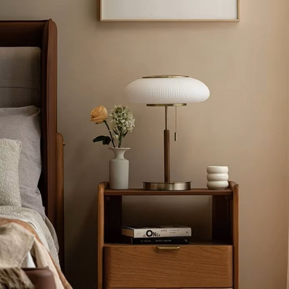A modern table lamp with a ribbed white oval shade and brushed brass base, styled on a wooden nightstand in a warm beige bedroom setting.