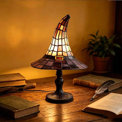 A stained-glass-style table lamp shaped like a witch hat sits on a wooden desk, glowing warmly. The lamp has a dark metal base and a conical shade made of amber, orange, and brown glass panels.