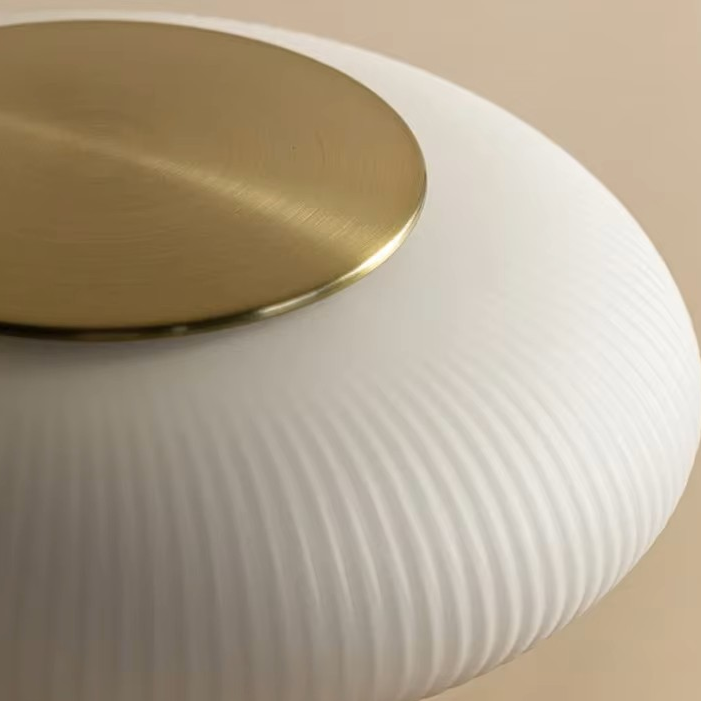 Close-up of a modern pendant light fixture with a ribbed white glass shade and a brushed gold metal top canopy, set against a warm beige background.