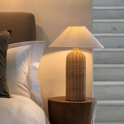 A bedside table lamp with a woven rattan-style textured base and a tapered off-white fabric shade, illuminated and placed on a rustic wooden nightstand next to a bed.