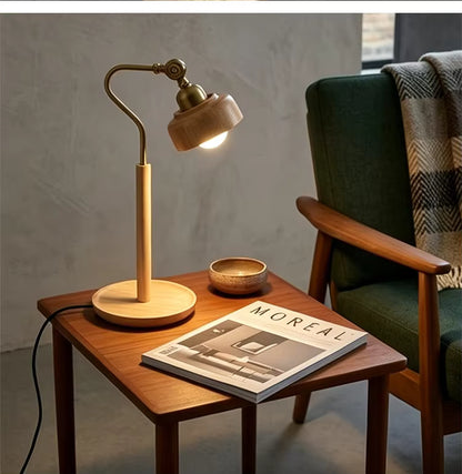 A mid-century modern table lamp with a light wood shade and base, mounted on a wooden side table next to a green armchair with a plaid throw blanket. The lamp is turned on, casting a warm glow, with a magazine and a small bowl on the table.